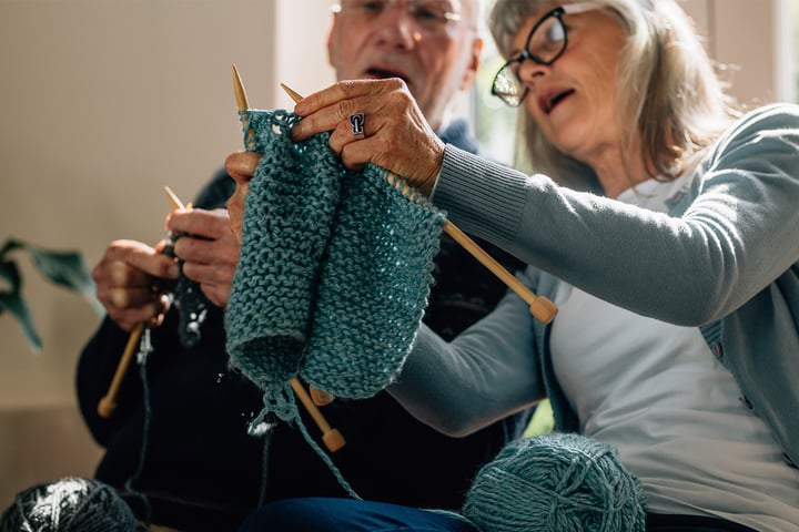 Older woman in glasses holds knitting up as she teaches older man