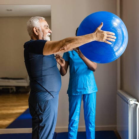 Senior man holds blue exercise ball in front of him with fitness instructor in background