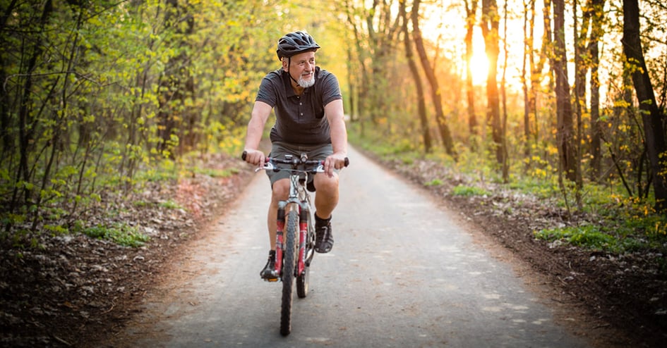 Senior man rides bike on forest path with sun rising in background