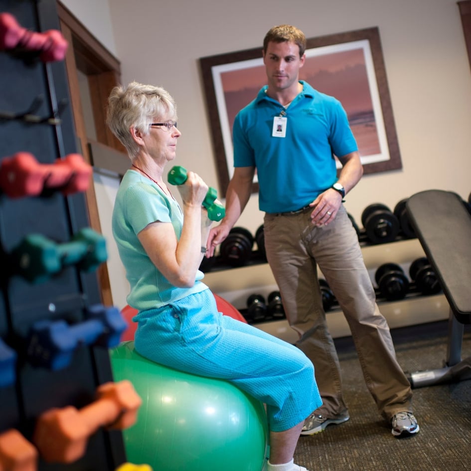 Senior woman holds weights and sits on balance ball in fitness room