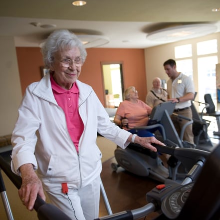 Senior woman in jogging suit talks on treadmill in busy fitness center