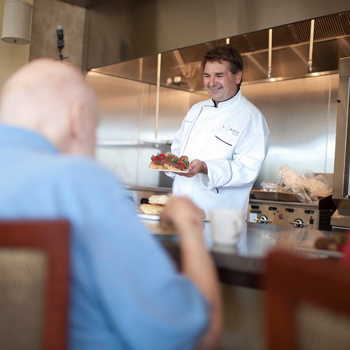 Chef in white coat presenting plated meal to elderly man sitting at bar